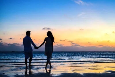 Мужчина и женщина, держась за руки, гуляют по берегу моря во время восхода  солнца · Бесплатные стоковые фото